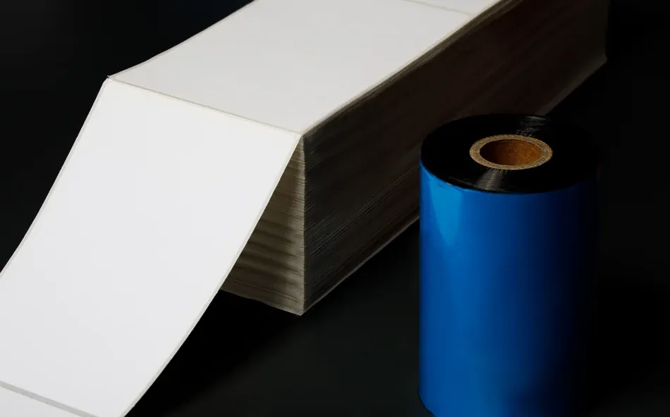 a stack of blank fanfold labels next to a roll of thermal transfer ribbon