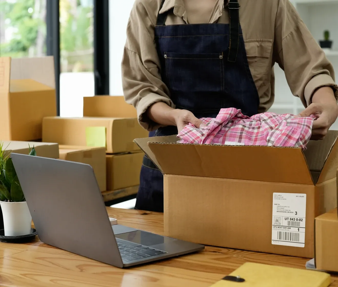 Person packing retail items in cardboard boxes with laptop and shipping labels on wooden desk